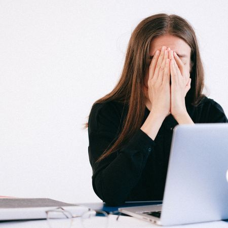 Zestresowana kobieta na jasnym tle pokoju siedząca przy laptopie służbowym ze złożonymi jak do modlitwy rękami - Proresult